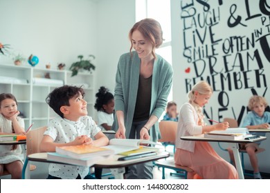Interacting with pupils. Cheerful pupil sitting at his desk and smiling to his kind teacher standing next to him. - Shutterstock ID 1448002802