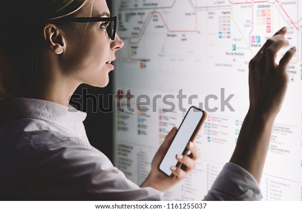 Interactive kiosk with public transport subway\
map.Female standing at big display with smartphone in hand.Young\
woman touching with finger screen while using train schedule\
application on mobile\
phone
