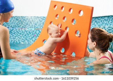 Interactive swimming training class with additional protective tools for infants. Little baby boy is using a floating mat with holes to get on his feet, mother and instructor is helping him