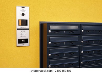 Intercom panel and modern dark mailboxes on the
vibrant yellow wall of apartment building. An outdoor multi-apartment IP video intercom system with numerical keypad next to a group of dark grey metal mailboxes mounted on a bright yellow wall.