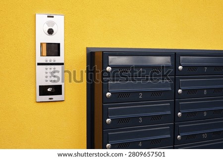 Intercom panel and modern dark mailboxes on the
vibrant yellow wall of apartment building. An outdoor multi-apartment IP video intercom system with numerical keypad next to a group of dark grey metal mailboxes mounted on a bright yellow wall.