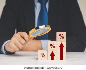 Interest rate financial concept. A Businessman is holding a magnifier and looking at wooden cubes with red arrows and percentage symbols on a wooden table.