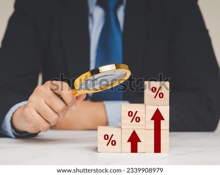 Interest rate financial concept. A Businessman is holding a magnifier and looking at wooden cubes with red arrows and percentage symbols on a wooden table.
