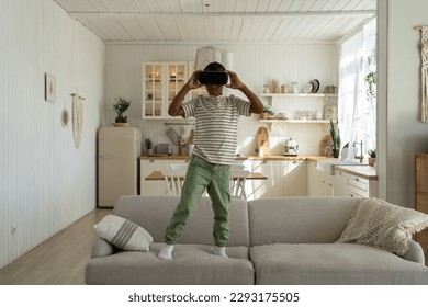 Interested african american boy plays video game stands on couch at home in virtual reality helmet. Interested preteen watches movie in vr goggles. Virtual reality, cyberspace, entertainment concept. - Powered by Shutterstock