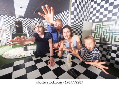 Interested Amazed Young Modern Family With Two Teen Sons Trying To Reach For Some Items In Escape Room Stylized As Chessboard, Holding Out Their Hands