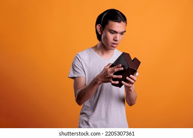 Interested Asian Man Opening Gift Box, Looking Inside Small Cardboard Parcel, Unpacking Surprise. Young Person Unboxing Present, Holding Delivery Package With Curious Facial Expression