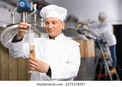 Interested brewery owner checking alcoholic content in craft beer, using test tube and hydrometer while standing near fermenters in workshop - Powered by Shutterstock