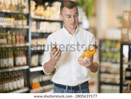 Interested casually dressed young man holding and examining organic juice bottles, pondering choices in natural food store
