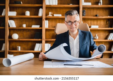 Interested caucasian mature middle-aged businessman engineer architect working on construction blueprints architectural plan, technical drawings in office looking at camera
