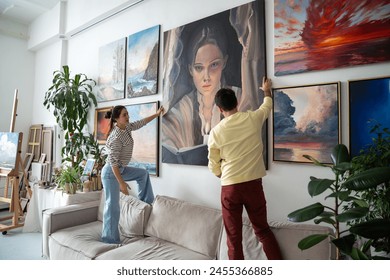 Interested couple of designers hang painting on wall looking on result talk about decoration standing on couch. Focused woman artist with man architect in home art studio discussion new art exhibition - Powered by Shutterstock