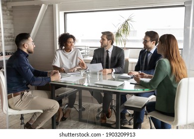 Interested diverse colleagues listening to leader, discussing statistics, sitting at table in modern boardroom, mentor coach explaining strategy, giving instructions to workers at briefing
