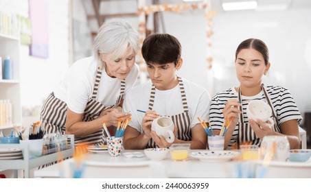 Interested elderly mother with wealth of pottery experience guiding young adult daughter and son working together on painting ceramic cups in family pottery workshop.. - Powered by Shutterstock