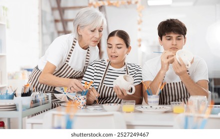 Interested elderly mother with wealth of pottery experience guiding young adult daughter and son working together on painting ceramic cups in family pottery workshop.. - Powered by Shutterstock