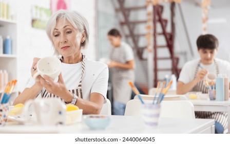 Interested elderly woman attending pottery class, enthusiastically painting on ceramic cup, creating unique custom design. Creative hobby concept - Powered by Shutterstock