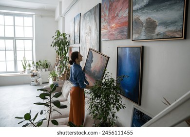 Interested female artist holds painting in hands and searches for suitable place on wall of home to hang result of creative work. Thoughtful designer woman decoration art studio with oil picture.  - Powered by Shutterstock