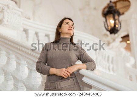 Interested female tourist admiring sumptuous interiors of antique palace while standing on stairs near white stucco railings, selective focus