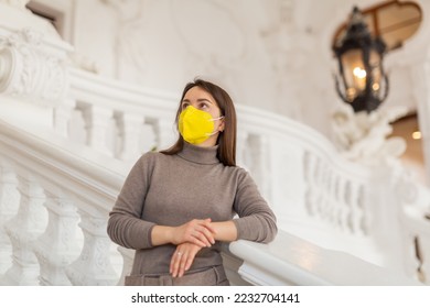 Interested female tourist in protective face mask admiring sumptuous interiors of antique palace while standing on stairs near white stucco railings. Covid pandemic precautions - Shutterstock ID 2232704141