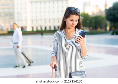 Interested Female Traveler Walking With Luggage Along Foreign City Streets, Reading About Sights In Smartphone..