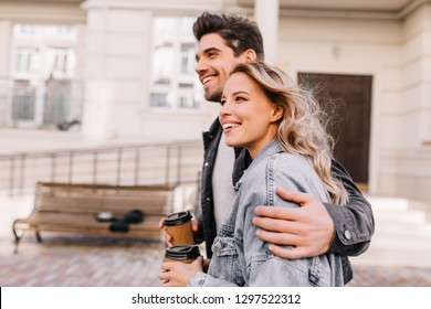 Interested girl drinking coffee during walk with boyfriend. Outdoor photo of happy young woman enjoying date.