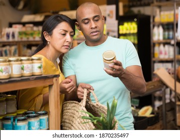 Interested Latin American couple reading product label on jar while choosing groceries in supermarket