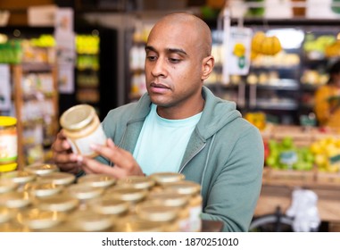 Interested Latin American Man Reading Product Label On Jar While Choosing Groceries In Supermarket