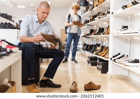 Interested man visiting shoe store in search of new shoes, choosing and trying on while sitting on pouf.