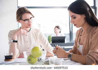 Interested Senior Female Employee Is Looking At Junior Colleague. She Glancing Into Notes And Telling Information