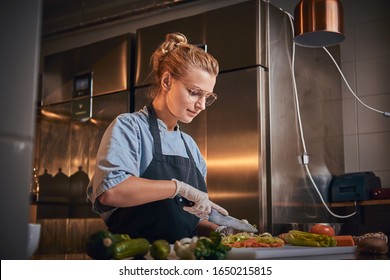 Interested and serious female chef standing in a dark restaurant kitchen next to cutting board while cutting vegetables on it, wearing apron and denim shirt, posing for the camera, cooking show look