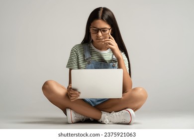 Interested student teen girl in glasses watching lection class on laptop on gray background. Focused teenager busy learning studies, study online. Education in college high school university concept.