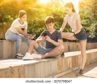 Interested Teen Boy Sitting With Phone On Summer City Street With Two Girls Trying To Get His Attention. Concept Of Phubbing Behavior