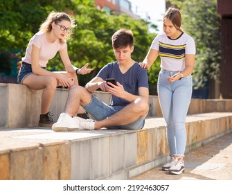 Interested Teen Boy Sitting With Phone On Summer City Street With Two Girls Trying To Get His Attention. Concept Of Phubbing Behavior