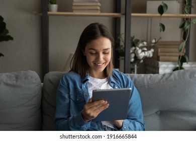 Interested teen female sit on cozy sofa spend time for reading electronic version of popular book at web library using reader app on tablet. Focused young lady write message email scroll data on pad - Shutterstock ID 1916665844