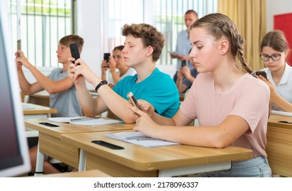 Interested Teen Girl Student Using Smartphone While Studying In College With Group Of Classmates..