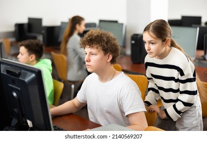 Interested Teen Schoolgirl Talking To Diligent Focused Classmate Sitting At Computer In Classroom During Informatics Lesson..