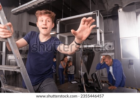 Interested teenage boy standing on stepladder reaching for something in closed quest room stylised as abandoned bunker while playing with his family