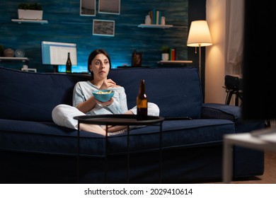 Interested Woman Eating Popcorn And Watching Entertainment Serial On Tv. Focused Home Alone At Night With Surprised Face Looking At Suspense Movie Sitting On Comfortable Interested Couch.