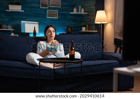 Interested woman eating popcorn and watching entertainment serial on tv. Focused home alone at night with surprised face looking at suspense movie sitting on comfortable interested couch.