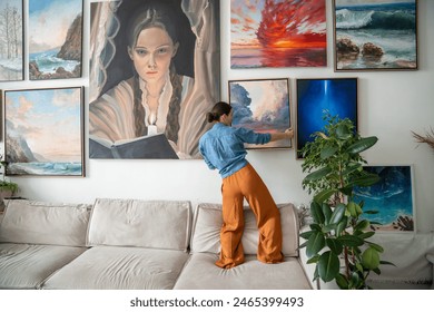 Interested woman painter, hangs on wall trendy artwork, created handmade in art workshop. Inspired pensive female artist designer holds new finished painting ponder compose for decoration art studio.