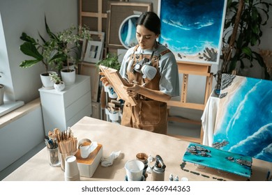 Interested woman painter holding wooden board in hands preparing for epoxy resin application standing in art studio. Small creative business. Focused artist girl in apron surround by pictures of sea. - Powered by Shutterstock