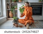 Interested woman takes care of home seedlings sits on kitchen floor, cuts leaves of green basil herbs. Female gardener pays attention to care of houseplants, tends, prunes excess and harvests crop.