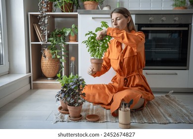 Interested woman takes care of home seedlings sits on kitchen floor, cuts leaves of green basil herbs. Female gardener pays attention to care of houseplants, tends, prunes excess and harvests crop. - Powered by Shutterstock