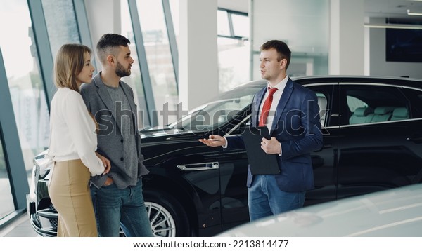 Interested young\
couple man and woman are talking to manager in car dealership while\
he is telling them about luxury auto model. Beautiful people,\
transportation and business\
concept.
