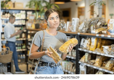 Interested young female shopper holding two different types of pasta, contemplating choice in well-stocked organic grocery store aisle - Powered by Shutterstock