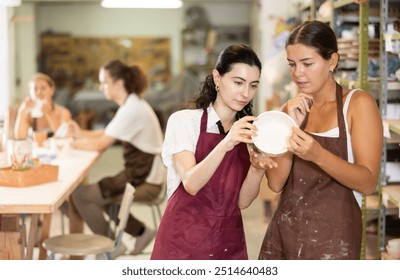 Interested young girls wearing aprons viewing attentively handmade clay bowl in pottery workshop - Powered by Shutterstock