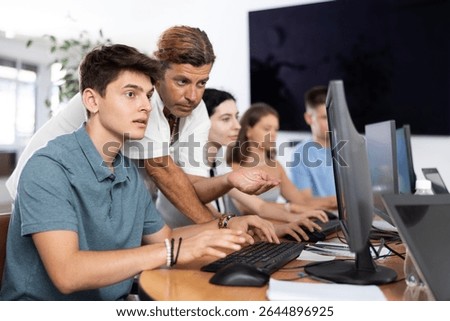 Interested young guy attending computer class, learning digital technology under guidance of positive adult male teacher standing nearby..