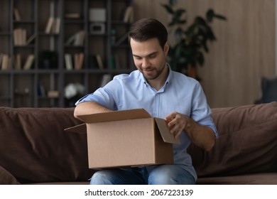 Interested Young Man Unpacking Big Carton Box, Sitting On Sofa At Home, Feeling Excited Of Getting Wished Items From Internet Store, Satisfied With Fast Delivery, Positive Online Shopping Experience.