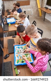 Interesting Building Kit For Kids On Table With Computers. From Above View Of Boys And Girls Creating Toys. Science Engineering. Nice Interested Friends Chatting And Working On Project Together.