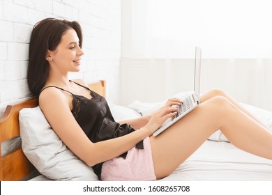 Interesting online news. Young girl reading blog on laptop, lying in bed, side view, empty space