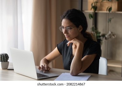 Interesting task. Focused young hispanic woman in eyewear sit by desk at home work online by laptop pc. Millennial female office worker employee search information at corporate network read e document - Shutterstock ID 2082214867