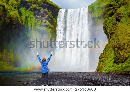 Interesting waterfall in Iceland - Skogafoss. Picturesque huge rainbow appears in the water mist. Middle-aged woman - tourist shocked beauty waterfall